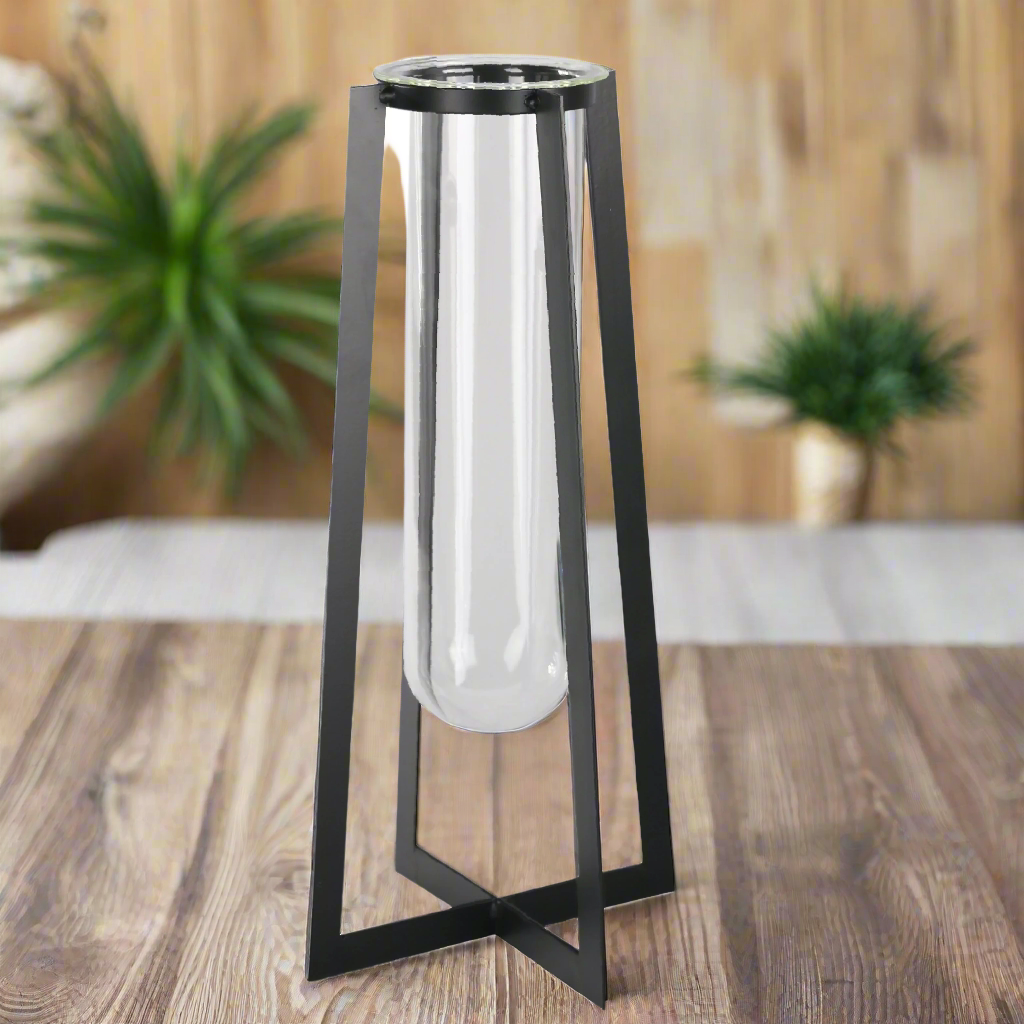 Black metal and glass cylindrical object on a wooden surface with plants in the background
