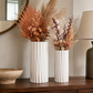 Two white ribbed ceramic vases filled with dried botanicals, displayed on a dark wood console table in a styled interior setting.