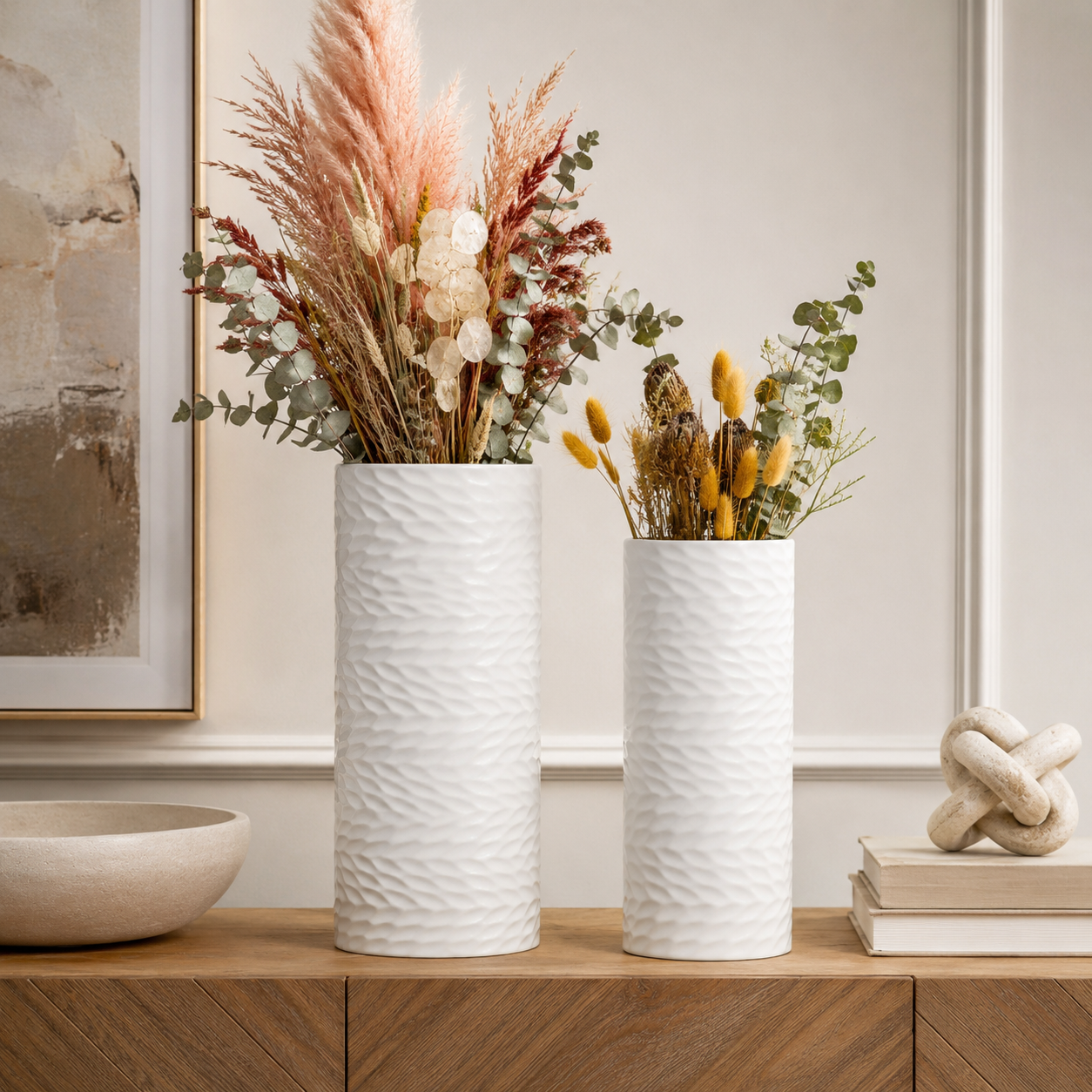 Two textured white ceramic vases filled with dried botanicals, displayed on a wooden console table in a styled interior setting.