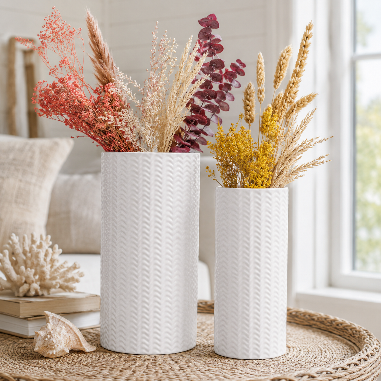 Two white textured ceramic vases filled with dried decorative grasses and flowers, displayed on a woven tray in a bright living room setting.
