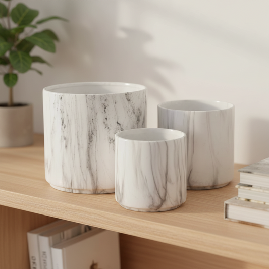 Three cylindrical planters in a white marble pattern with gray veining, displayed on a light wood shelf next to a potted plant and books.