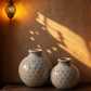 Two ceramic vases on a wooden shelf with warm lighting and shadows on a textured wall.