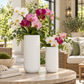 Two textured white ceramic flower vases filled with white, pink, and magenta orchids and green foliage, displayed on a round stone table in a bright, modern outdoor patio setting.
