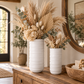 Two white ribbed ceramic vases filled with dried pampas grass, eucalyptus, and cream flowers, displayed on a wooden console table in a warm, styled interior.