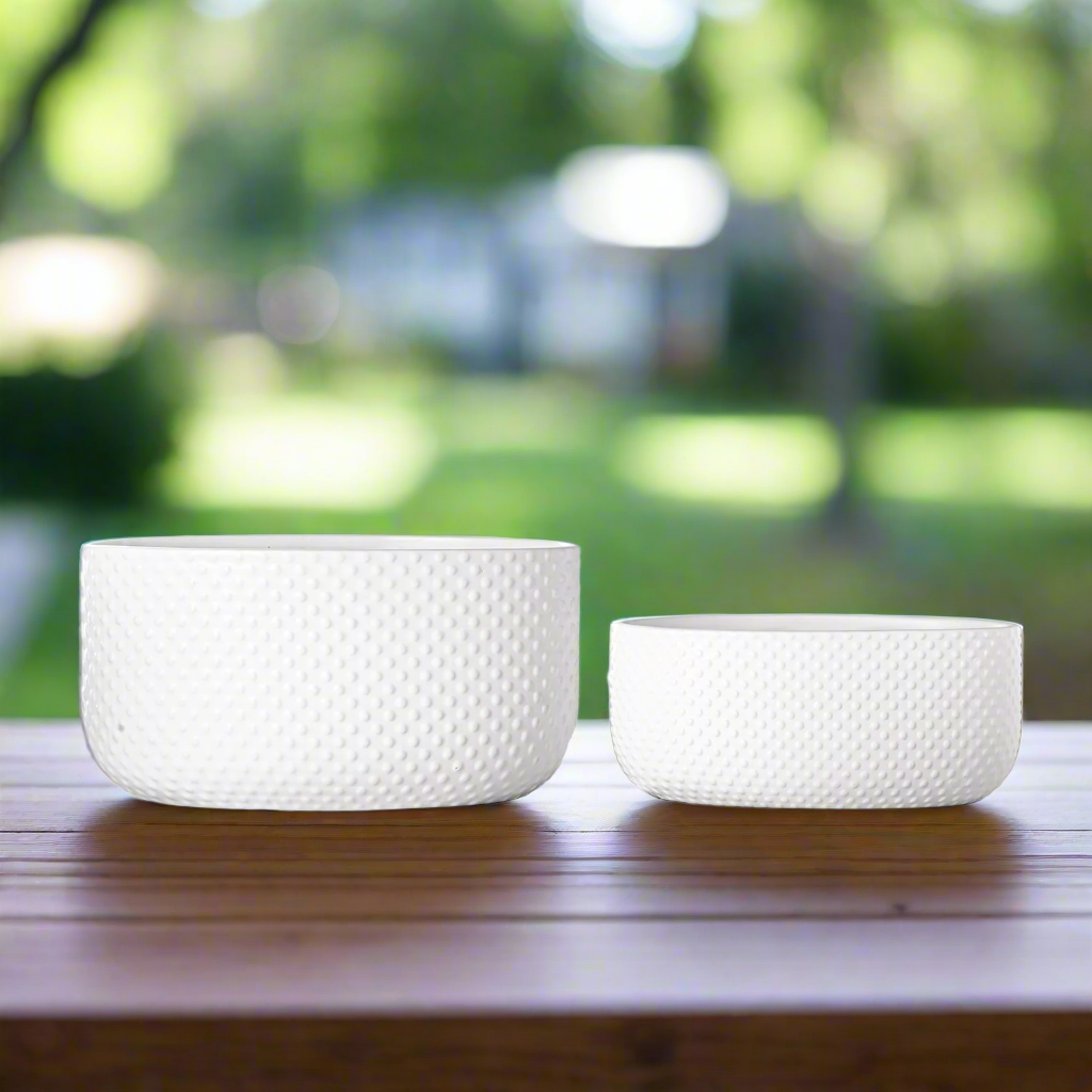 Two white textured planters on a wooden surface with a blurred green outdoor background