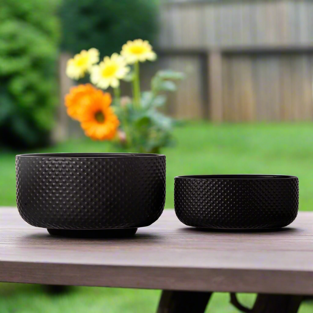 Two black textured planters on a wooden table with flowers in the background.