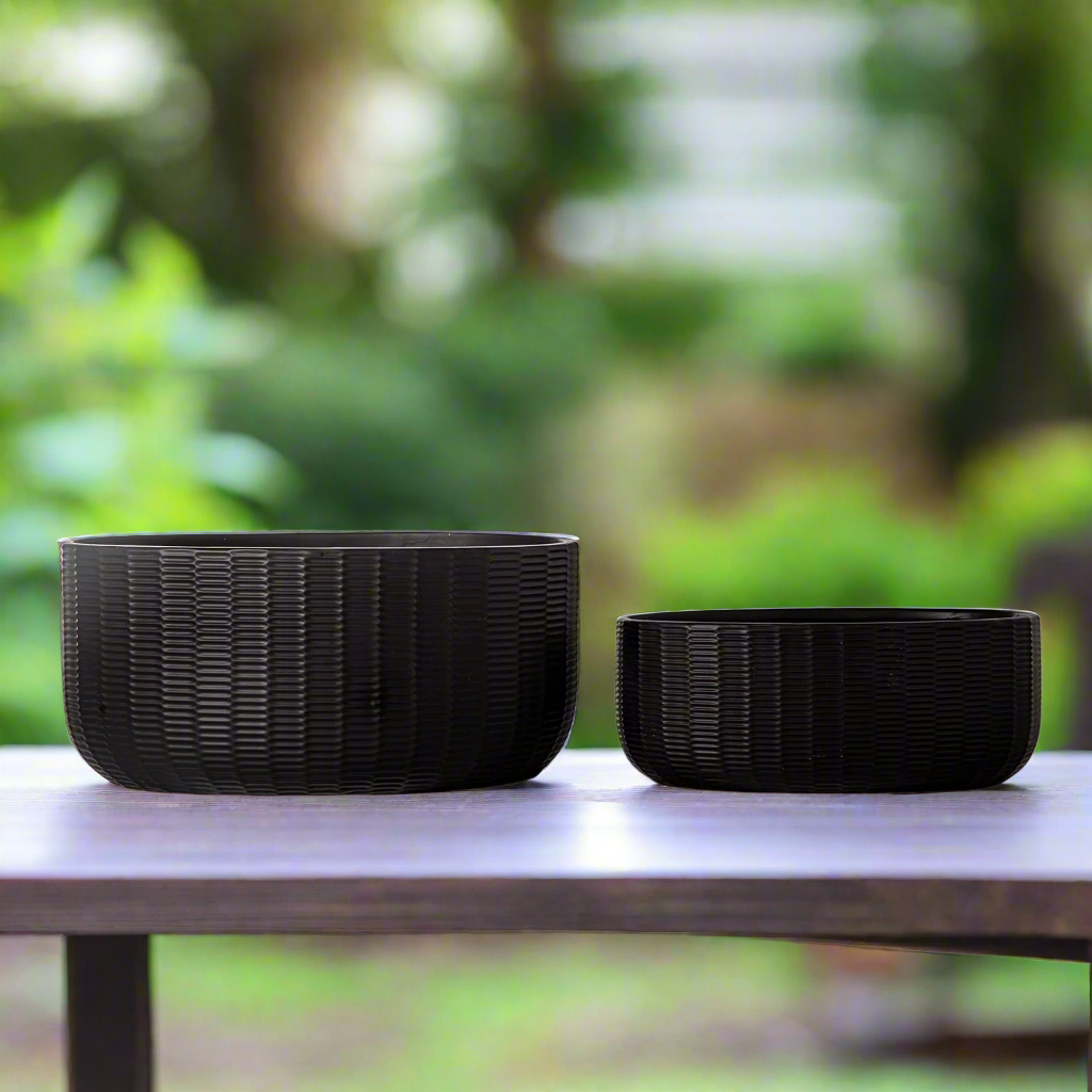 Two black woven bowls on a wooden table with a blurred green outdoor background
