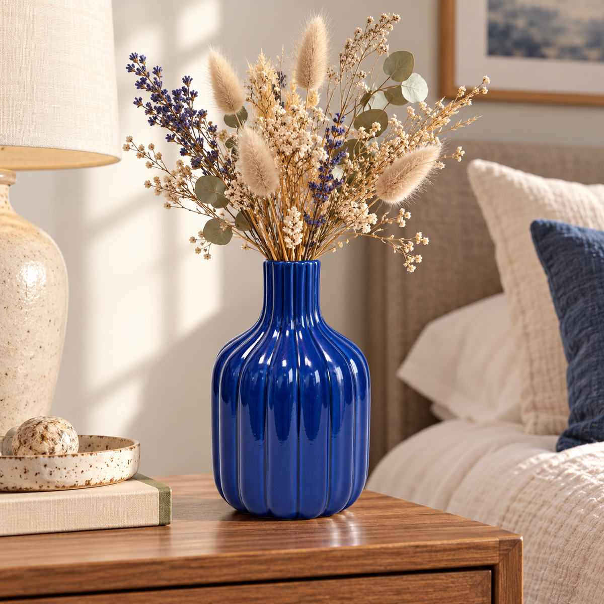 A glossy cobalt blue ribbed ceramic vase filled with dried grasses, eucalyptus leaves, and small purple flowers, styled on a wooden nightstand in a bedroom setting.