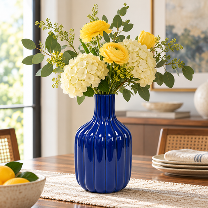 A glossy cobalt blue ribbed ceramic vase filled with yellow ranunculus and cream hydrangeas, styled on a woven placemat on a wooden dining table in a bright, modern room.