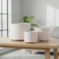 Three round off-white ceramic planters with a subtle woodgrain texture, displayed in a nested size progression on a light wooden table in a bright, modern room.
