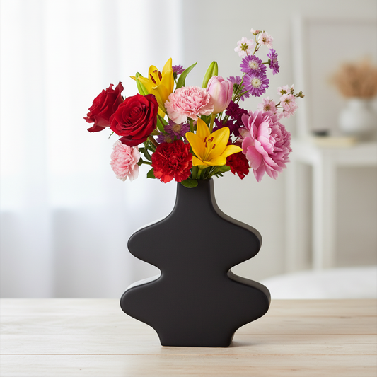 A black sculptural ceramic vase filled with a colorful bouquet of red, pink, yellow, and purple flowers, set on a light wood tabletop with a softly blurred indoor background.