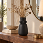 A matte black ceramic vase with a sculptural, rounded form and small protruding side details, filled with dried beige flowers, displayed on a dark wood console table in a styled interior setting.