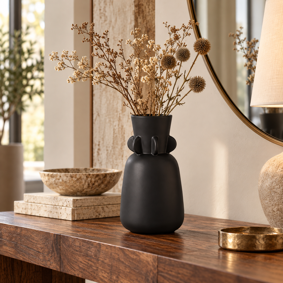 A matte black ceramic vase with a sculptural, rounded form and small protruding side details, filled with dried beige flowers, displayed on a dark wood console table in a styled interior setting.