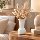 A white ceramic vase filled with dried beige grasses and small cream flowers, styled on a wooden bedside table next to a lamp in a softly lit bedroom setting.
