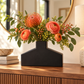 An artificial floral arrangement with orange protea-like flowers, peach ranunculus, green leaves, and small berry clusters in a matte black geometric vase, displayed on a wooden console table in a warmly lit interior.