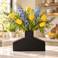 A black ceramic vase with a modern house-shaped base, filled with a spring floral arrangement of yellow tulips, blue delphiniums, and green eucalyptus, set on a white marble kitchen countertop.