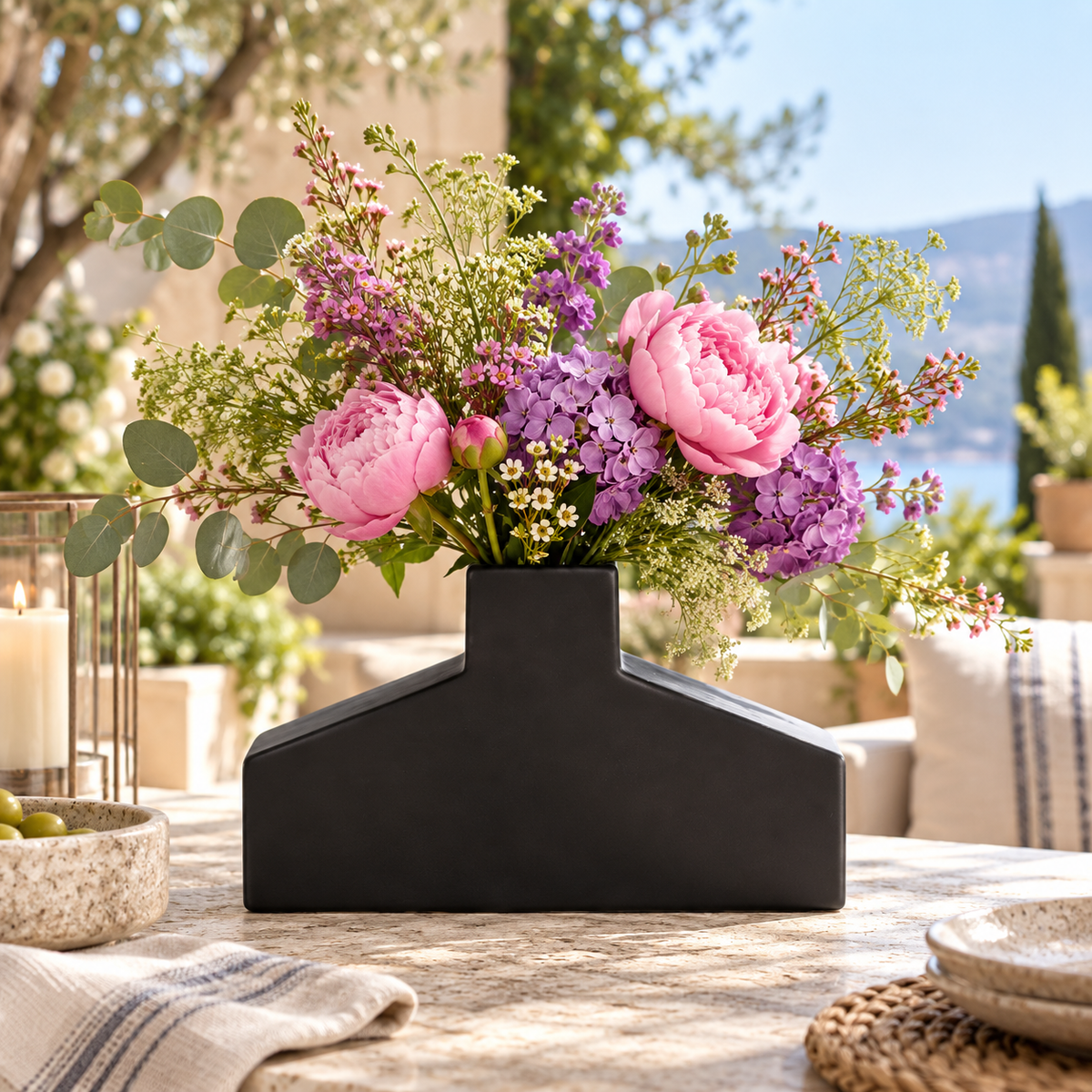 A black geometric ceramic vase filled with a colorful wildflower arrangement featuring pink peonies, purple flowers, and green eucalyptus, set on a rustic outdoor patio table.