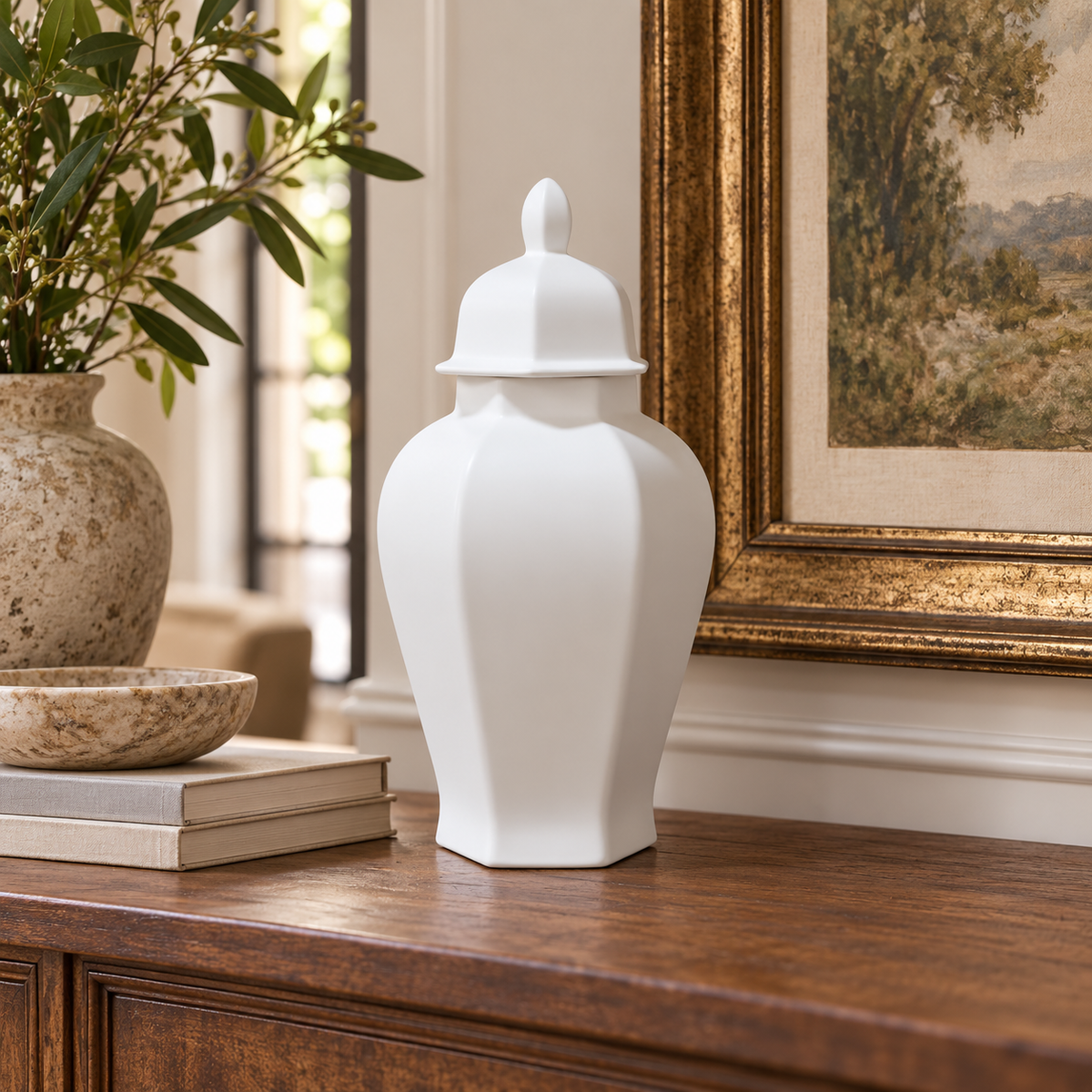 A white ceramic ginger jar with a lid sits on a dark wood console table in a styled interior setting next to a framed painting and a potted plant.