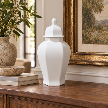 A white ceramic ginger jar with a lid sits on a dark wood console table in a styled interior setting next to a framed painting and a potted plant.