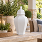 A white ceramic lidded urn with a geometric faceted design sits on a stone patio table next to a woven tray, candle lantern, and potted green plants.