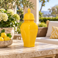 A glossy bright yellow ceramic lidded jar vase sits on a stone table in an outdoor patio setting, with a potted plant of white flowers and lemons on the left and a wicker sofa in the background.