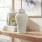 A white ribbed ceramic lidded vase with a pointed finial, displayed on a light wood shelf next to stacked books and a small succulent.