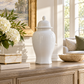 A glossy white ceramic lidded jar with a rounded body and small finial top, displayed on a wooden console table in a bright, sunlit living room setting.