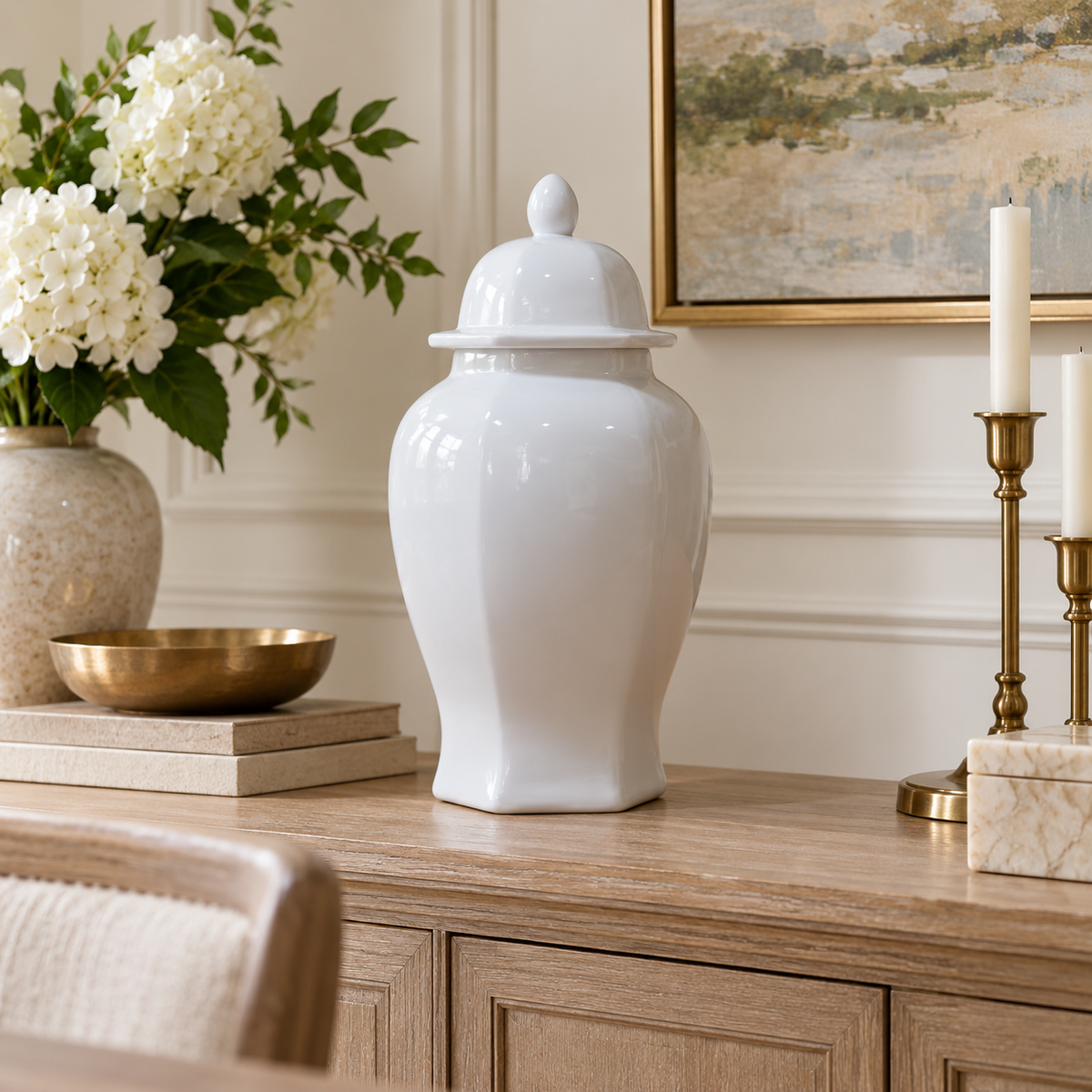 A glossy white ceramic ginger jar with a matching lid sits on a light wood console table in a styled room setting, surrounded by a vase of white flowers, gold bowls, and brass candlesticks.