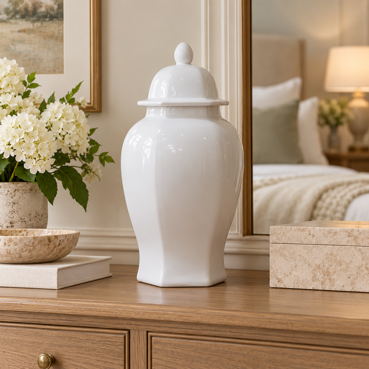 A glossy white ceramic lidded jar or canister sits on a wooden dresser in a softly lit bedroom setting, with flowers, books, a mirror, and bedding visible in the background.