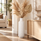 A tall white textured ceramic vase filled with beige dried grasses and palm leaves, placed in a bright, modern living room next to a light wood cabinet.