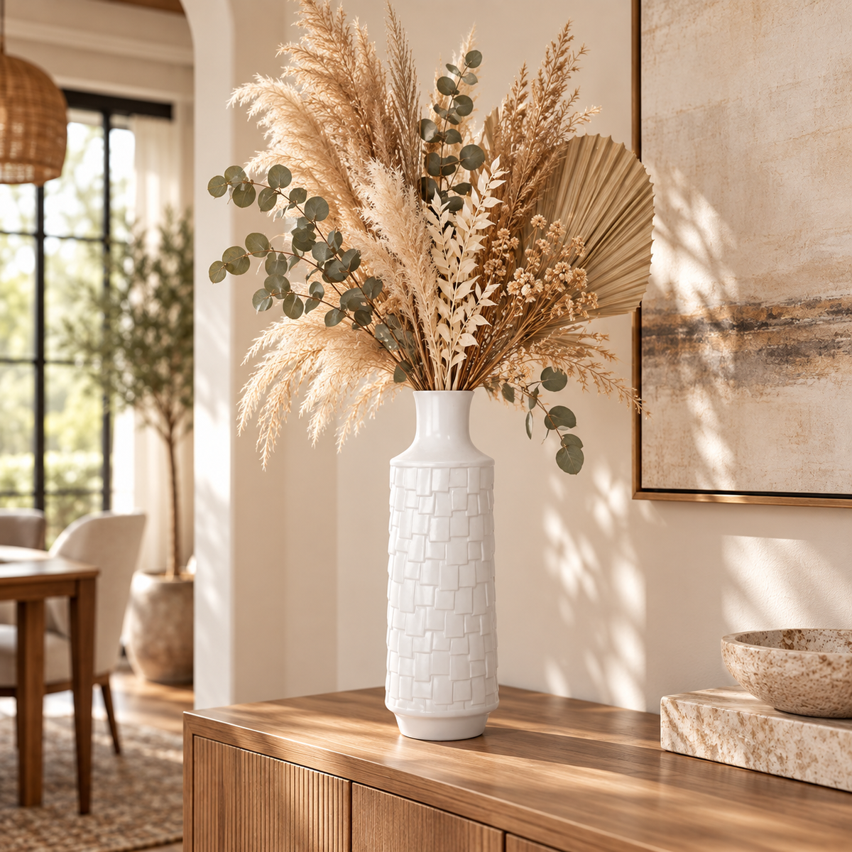 A tall white textured ceramic vase filled with dried pampas grass, eucalyptus leaves, and a beige palm fan, displayed on a wooden console table in a warm, modern living room.