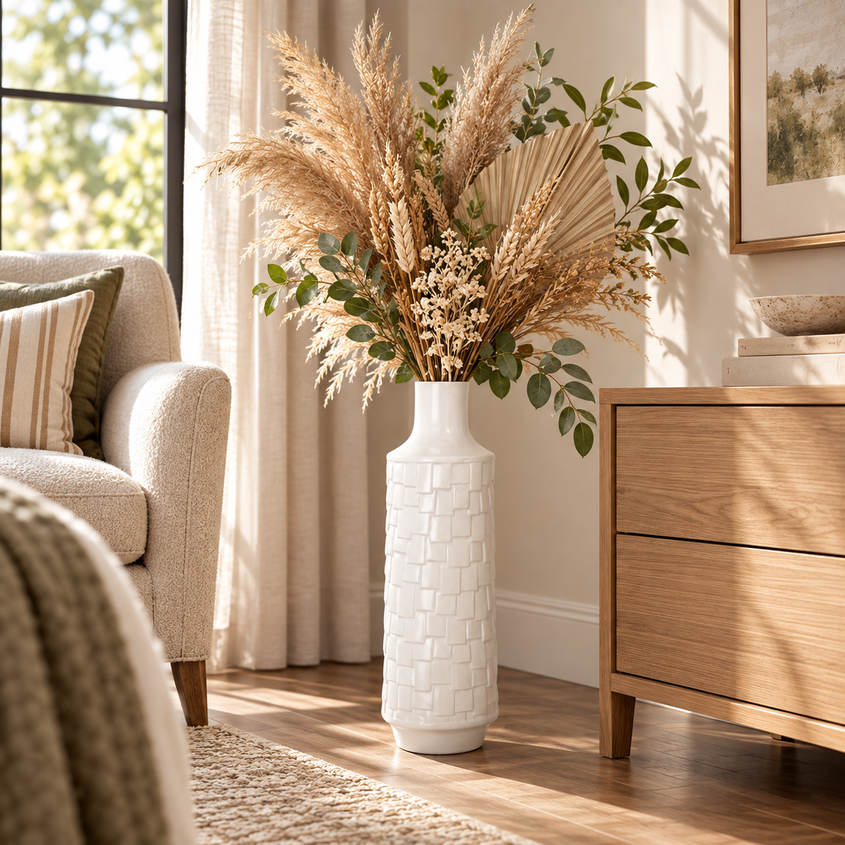 A tall white textured ceramic vase filled with dried pampas grass, wheat, and green foliage, styled in a bright, modern living room next to a wooden dresser and armchair.