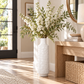 A tall white textured ceramic vase filled with white blossoms and green foliage, styled on a light wood floor next to a wooden console table in a bright, modern room.