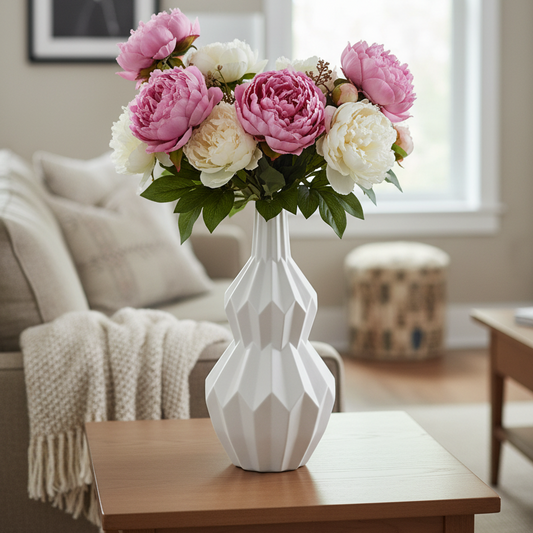 A white geometric ceramic vase filled with pink and cream peony flowers, placed on a wooden side table in a softly lit living room.