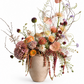 A rustic beige ceramic vase filled with a lush, multi-colored wildflower arrangement featuring orange ranunculus, pink anthuriums, purple globe flowers, and delicate trailing greenery, set against a solid white background.
