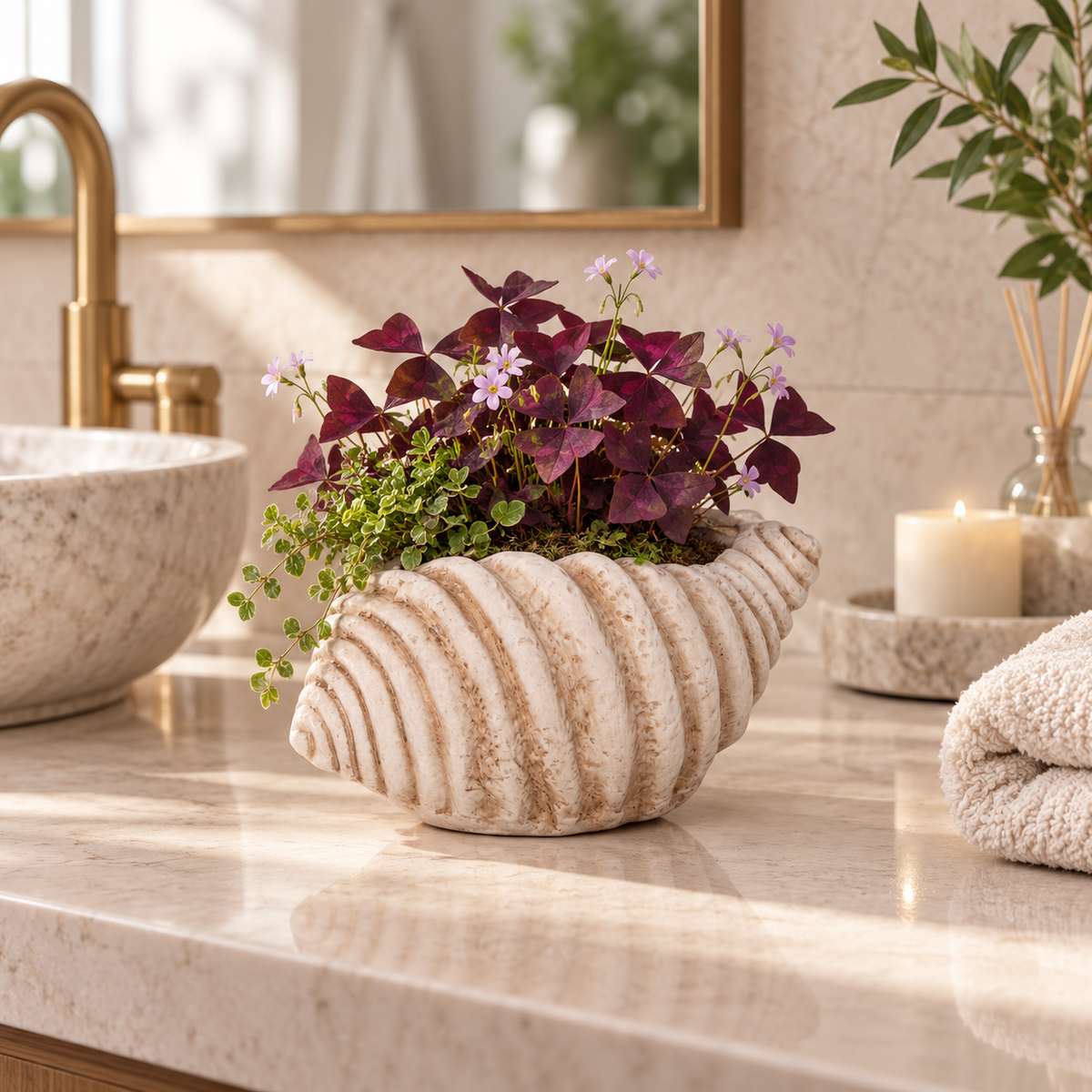 A seashell-shaped planter with a textured cream and beige finish, filled with green and burgundy foliage and small purple flowers, set on a marble bathroom counter next to a gold faucet and a lit candle.