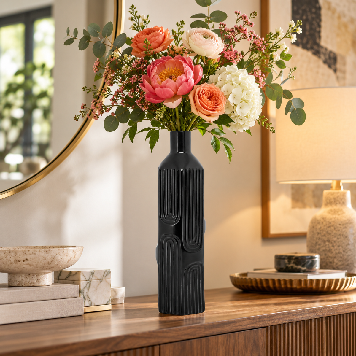 A black ribbed ceramic vase holding a colorful floral arrangement with pink, peach, white, and green flowers, displayed on a wooden console table in a warmly lit interior setting.