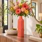 A tall ribbed coral-orange ceramic vase filled with a colorful floral arrangement, displayed on a wooden console table in a bright, modern room.