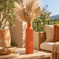 An orange terracotta ribbed ceramic vase filled with dried pampas grass and decorative stems, styled on a wooden table in a sunny outdoor patio setting with a woven chair and plants in the background.