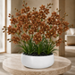 A potted artificial orchid arrangement with many medium-brown flowers, green leaves, and black stones in a round white planter, placed on a round stone table in a softly lit interior setting.