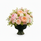 A floral arrangement of pink and peach roses, peonies, white blossoms, and green foliage in a dark green pedestal urn, photographed on a clean white background.