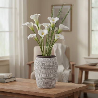 A decorative gray ceramic vase with a textured geometric pattern, filled with white calla lilies and green stems, placed on a wooden table in a softly lit living room.