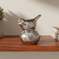 A polished silver metal decorative sculpture shaped like a bird perched on a rounded base, displayed on a wooden shelf against a warm beige wall.