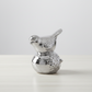 A polished silver bird figurine perched on a textured round base, photographed on a light tabletop against a soft light gray background.