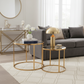 Two round nesting side tables with gold metal frames and clear glass tops in a modern living room setting, styled with a brass table lamp, books, and a vase of white flowers in front of a gray sofa.
