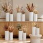 A styled interior scene showing multiple white textured ceramic vases filled with dried grasses and botanicals, arranged on a wooden cabinet and floor against a warm neutral wall.