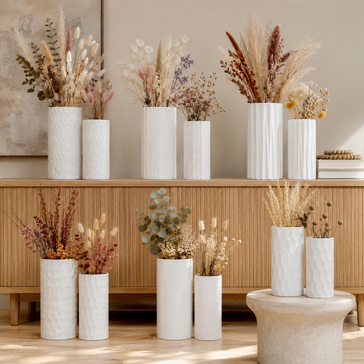 A styled interior scene showing multiple white textured ceramic vases filled with dried grasses and botanicals, arranged on a wooden cabinet and floor against a warm neutral wall.
