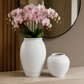 Two white textured ceramic vases, one tall with pink orchids and one short, displayed on a light stone console table in a modern interior setting.