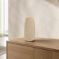 A tall, textured cream-colored ceramic vase sits on a light wood sideboard in a modern, minimalist room with warm neutral tones.