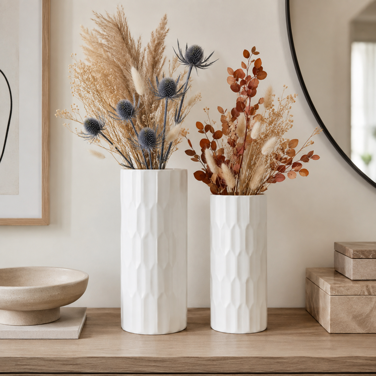 Two textured white ceramic vases filled with dried decorative grasses and botanicals, displayed on a wooden console table against a warm beige wall.
